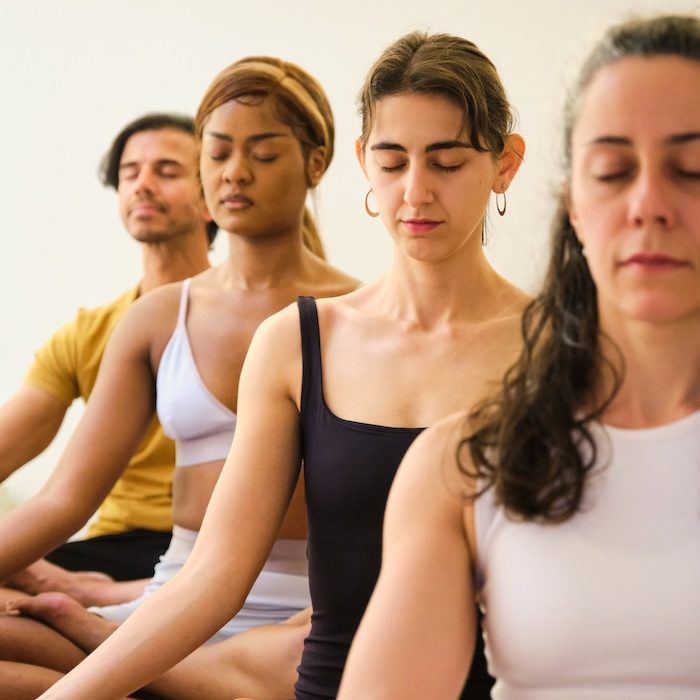 A group of people are sitting on yoga mats and meditating. Scene is peaceful and serene, as the people are meditating and focusing on their breathing.