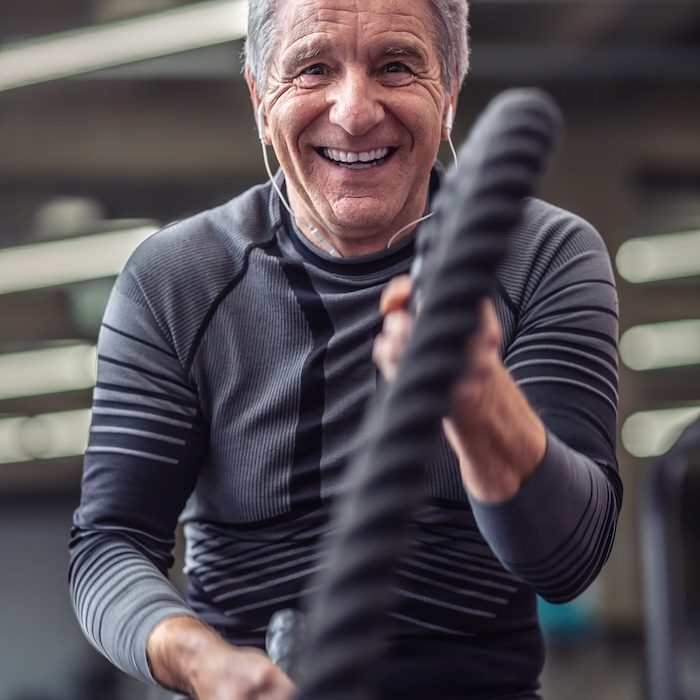 Older man laughs while working out with ropes inside the gym, wearing headphones.